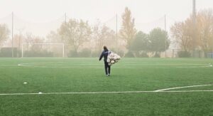 A person with a net full of soccer balls walking down a soccar field with the goal in the distance