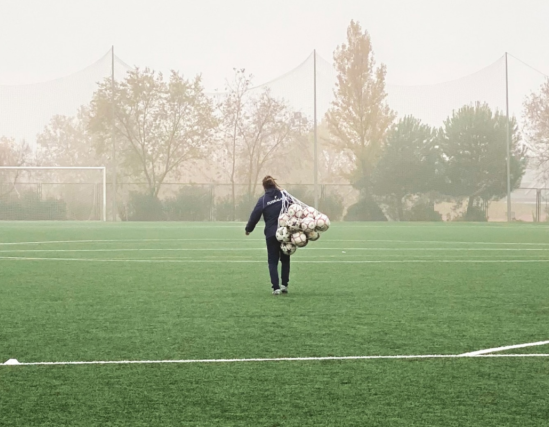 A person with a net full of soccer balls walking down a soccar field with the goal in the distance