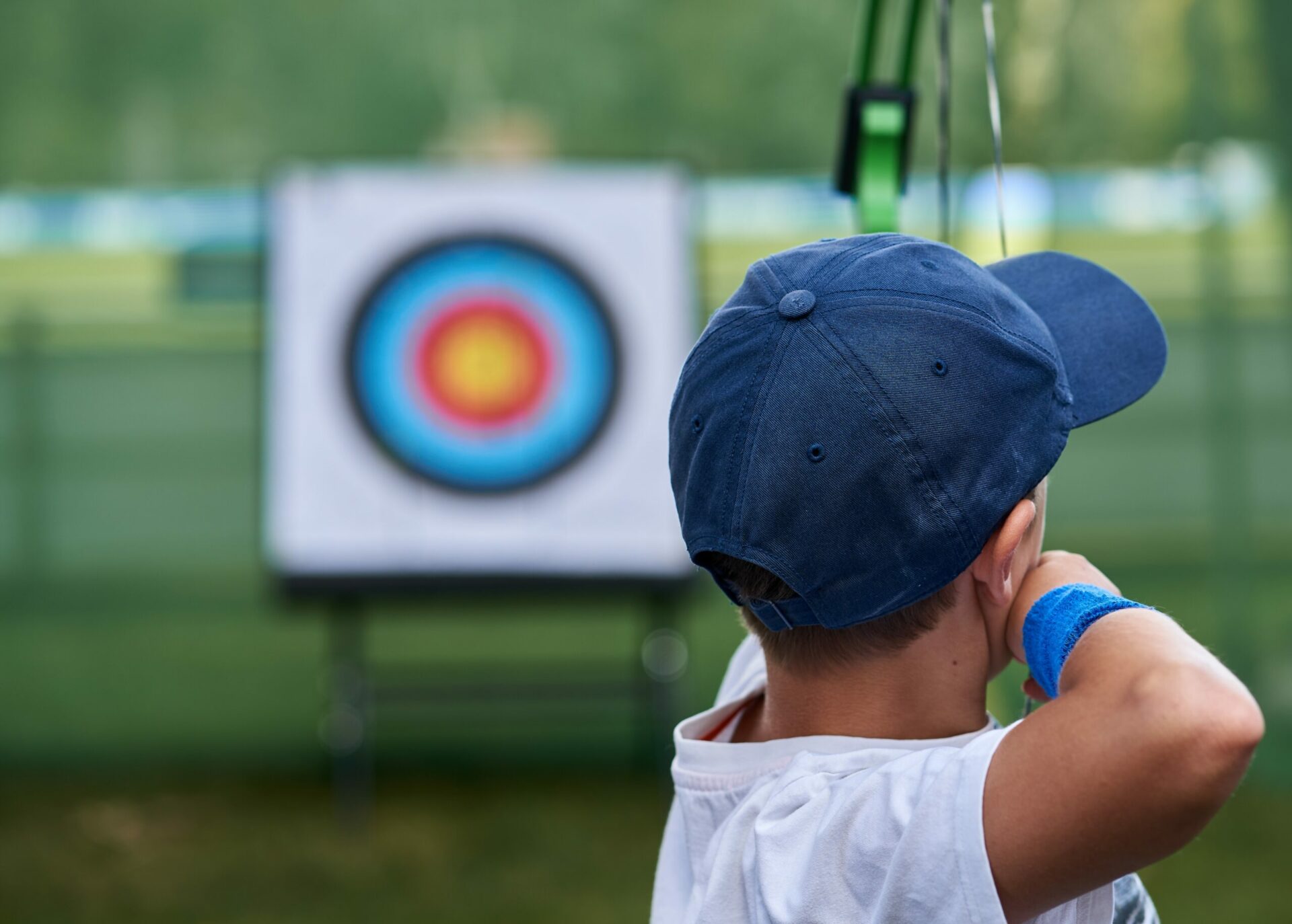 Photo from behind of a boy wearing a blue cap pulling back his bow to shoot an arrow at a target in the distance.